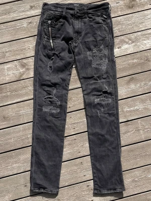 Replay Anbass Distressed Slim Jeans – W31 L32 Svart/Grå - Säljer ett par Replay Anbass jeans i slim fit.  Svart/mörkgrå tvätt med kraftiga slitsningar och destruktioner framtill.  Har även en dragkedjedetalj på låret.  Storlek: W31 L32  Skick: Bra begagnat skick. Tvättade och klara. Slitsningarna är en del av designen.  Perfekt för grisch/streetwear-look.  Mått kan tas vid förfrågan.