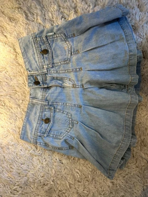 Ljusblåa högmidjade jeansshorts - Säljer ett par ljusblåa högmidjade jeansshorts med två framfickor och dekorativa knappar. Shortsen har en lös passform och är tillverkade i mjuk denim. Perfekta för varma dagar och enkel att matcha med olika toppar.