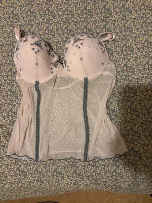 Vit mesh body med blå broderi - Söt vit body i mesh med vadderade kupor och grått broderi samt små rosa blommor. Transparent tyg med prickig struktur och dekorativa blå sömmar framtill. Axelbanden är justerbara och materialet känns lätt och luftigt.