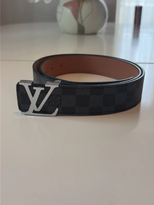 rutigt svart bälte - Snyggt svart bälte från Ł V med klassiskt rutigt mönster och stor Ł V-logga i silver på spännet. Bältet är gjort i skinn med brun insida och har en exklusiv känsla som lyfter vilken outfit som helst.