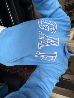 Blå GAP hoodie  - Snygg ljusblå hoodie från GAP med stor vit och blå logga över bröstet. Klassisk känguruficka framtill och mjukt material som känns skönt mot huden. Perfekt för en avslappnad och trendig look💙
