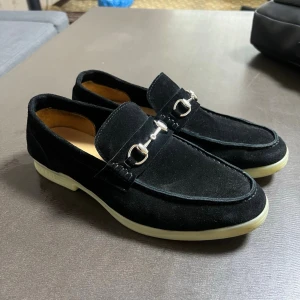 Svarta mocka loafers från Gucci - Snygga svarta loafers i mocka från Gucci med klassisk metallspänne framtill. Skorna har en stilren design med rundad tå och kontrasterande vit sula. Perfekta för dig som vill ha en tidlös och trendig look.