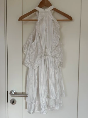 Vit omlottklänning med volanger - Säljer en vit kortklänning med tunna vertikala ränder och volangdetaljer. Klänningen har halterneck och omlottkänsla, vilket ger en luftig och somrig look. Perfekt för varma dagar och har en lätt, flowy passform.