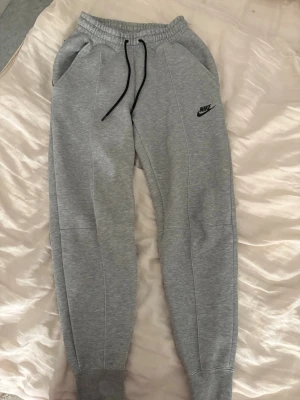 Gråa Nike tech mjukisbyxor från Nike - Snygga gråa mjukisbyxor från Nike med svart logga på benet. Byxorna har resår i midjan med svarta dragsnören, två sidofickor och ribbade muddar vid bensluten. Perfekta för chill dagar eller träning.