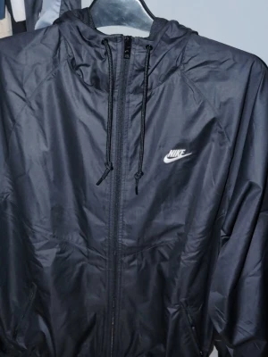 Svart vindjacka från Nike st M passar S också  - Snygg svart vindjacka från Nike med dragkedja framtill och justerbar huva med snören. Jackan har Nike-logga i vitt på bröstet och är tillverkad i ett lätt, vindtätt material. Perfekt för blåsiga dagar och enkel att matcha med olika outfits.HELT NY passar de mesta har exakt likadan typ så onödigt ha den liggandes ..ordinarie pris tusen ungefär säljes för 699