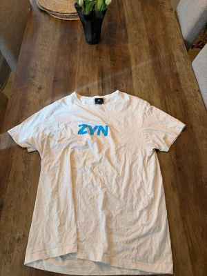 Vit ZYN t-shirt med blå text - Säljer en vit t-shirt med blå ZYN-tryck på bröstet. Klassisk rund hals och korta ärmar. Perfekt för dig som gillar enkel och clean stil. Tillverkad i mjuk bomull för skön känsla hela dagen.