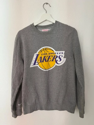 Grå Lakers sweatshirt Mitchell & Ness - Säljer en grå sweatshirt från Mitchell & Ness med stort Los Angeles Lakers-tryck på bröstet. Tröjan har rund halsringning och långa ärmar. Perfekt för dig som gillar basket och vill ha en sportig look.