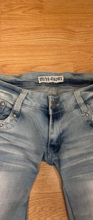 Ljusblå Miss Capri jeans med detaljer - Säljer ett par ljusblå jeans från Miss Capri med slitningar och snygga dekorationer på fickorna. Jeansen har smal passform, silverfärgade nitar och ett broderat mönster på bakfickan. Perfekta för dig som gillar en unik och trendig stil.