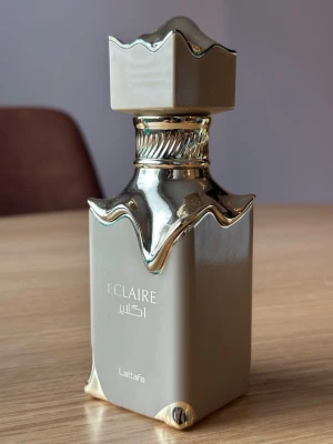 Eclaire parfym från Lattafa - Säljer en elegant parfymflaska från Lattafa, modellen Eclaire. Flaskan är fyrkantig i beige och guld med en unik, vågig design upptill och ett matchande lock. Text i både engelska och arabiska på flaskan. Perfekt för dig som gillar lyxig design.