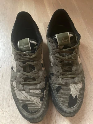 Valentino camo sneakers i grönt - Snygga sneakers från Valentino med camo-mönster i olika nyanser av grönt, beige och svart. Skorna har klassisk låg profil, gröna snören och ovandel i textil och mocka. Perfekta för dig som vill sticka ut med en unik streetstyle.