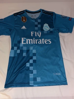 Blå Real Madrid matchtröja Adidas - Säljer en blå Real Madrid matchtröja från Adidas med CLIMACOOL-material. Tröjan har digitalt rutmönster, klubbmärke och sponsortryck framtill samt svarta ränder på axlarna. Perfekt för fotbollsträning eller att bära till vardags.