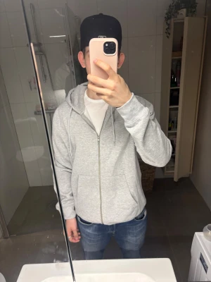 Ljusgrå zip hoodie med huva Lindbergh - Säljer en enkel och stilren ljusgrå hoodie med dragkedja och huva. Hoodien har fickor framtill och justerbara snören i huvan. Nypris 899kr. Den är helt oanvänd och har prislappen kvar. 