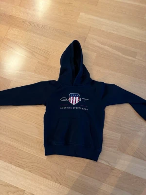 Mörkblå hoodie från GANT - Snygg mörkblå hoodie från GANT med klassisk huva och magficka. Framtill finns ett broderat GANT-emblem med amerikansk flaggsköld och texten 'American Sportswear'. Tillverkad i mjukt bomullsmaterial, perfekt för en avslappnad look.