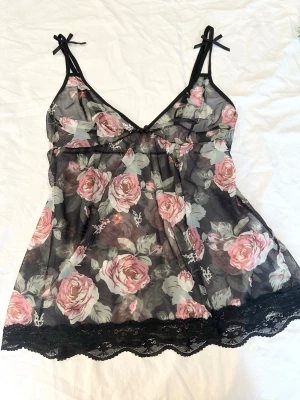 Blommig transparent linnetopp med spets - Säljer en romantisk och transparent linnetopp med rosa blommor på svart botten. Toppen har tunna axelband med små rosetter och spetskant nertill. Perfekt för dig som gillar en feminin och luftig stil.