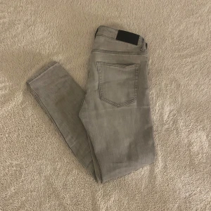Grå slim fit jeans från Dressmann - Säljer ett par grå slim fit jeans från Dressmann med klassisk femficksdesign och knappstängning. Jeansen har en snygg, ljusgrå tvätt och smal passform som sitter tajt längs benen. Perfekt för dig som gillar stilrena och moderna jeans. Om du köper skulle de uppskattas om du köper med PostNord eller instabox. Pris kan diskuteras 