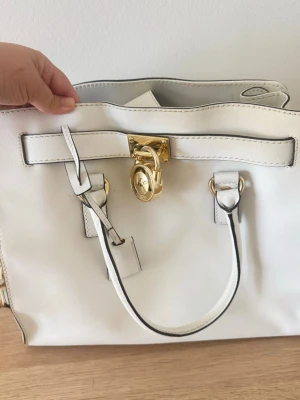 Michael Kors Hamilton väska - Stilren vit handväska i läderimitation med guldfärgat lås och detaljer. Väskan har två handtag och dekorativ rem framtill. Perfekt för dig som gillar klassisk och elegant design.
