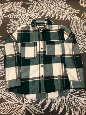 Grönrutig overshirt från Zara - Snygg overshirt från Zara i grönt, vitt och brunt rutmönster. Två stora bröstfickor med lock, knäppning framtill och klassisk krage. Perfekt lager på lager-plagg i mjukt, vävt material. Passar dig som gillar avslappnad och trendig stil.