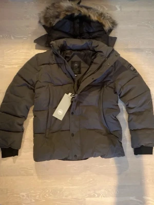 Svart dunjacka med pälshuva - Snygg svart dunjacka från Canada Goose med avtagbar huva och fuskpälsdetalj. Jackan har två sidofickor, ribbade muddar och klassisk patch på ärmen. Perfekt för kalla vinterdagar och riktigt varm tack vare dunfyllningen.