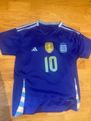 Argentina Messi matchtröja blå - Snygg blå Argentina matchtröja från Adidas med nummer 10 och Messi på ryggen. Tröjan har FIFA-märke på bröstet, ljusblå detaljer och klassiska Adidas-ränder på axlarna. Tillverkad i lätt och ventilerande material, perfekt för fotboll eller träning.