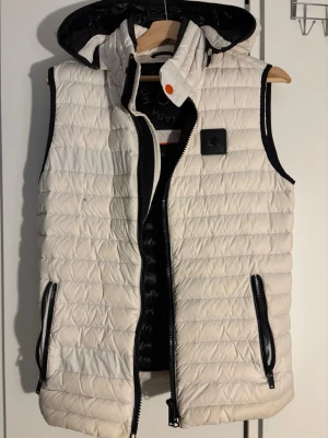 Vit dunväst från Moose Knuckles - Snygg vit dunväst från Moose Knuckles med svart avtagbar huva och svarta detaljer. Västen har två dragkedjeförsedda fickor, quiltad design och en diskret logga på bröstet. Perfekt för lager-på-lager och håller dig varm med stil. Och passar även s 