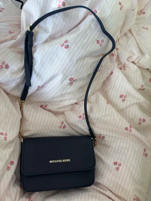 Mörkblå axelväska Michael Kors - Snygg mörkblå axelväska från Michael Kors i stilren design. Väskan har justerbar axelrem, guldfärgade detaljer och är gjord i ett tåligt, strukturerat konstläder. Perfekt storlek för det viktigaste och enkel att matcha till olika outfits.