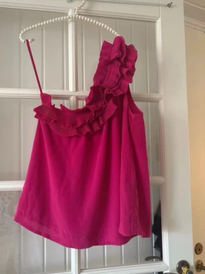 Rosa oneshoulder topp med volang - Säljer en snygg rosa oneshoulder topp med bred volangdetalj längs halsringningen. Toppen är ärmlös och har en cool, asymmetrisk design som sticker ut. Perfekt för dig som vill ha något unikt och trendigt i garderoben.