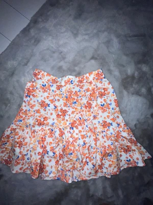 Blommig volangkjol från Lindex XS - Supersöt kort kjol från Lindex i vitt med orange, blått och gult blommönster. Kjolen har volangdetaljer och omlottkänsla, perfekt för en fräsch och somrig look. Resår i midjan för extra komfort.