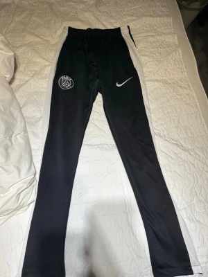 Svarta PSG träningsbyxor Nike - Snygga svarta träningsbyxor från Nike med Paris Saint-Germain-logga på ena benet och vit Nike-swoosh på andra. Byxorna har vita sidopaneler och smal passform, perfekta för fotboll eller gym. Tillverkade i ett stretchigt funktionsmaterial.