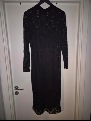 Svart lång spetsklänning från H&M  - Elegant svart långklänning i spets med hög hals och långa ärmar. Klänningen har ett vackert blommigt spetsmönster som ger en exklusiv känsla. Perfekt för dig som vill ha en stilren och sofistikerad look. Använd en gång
