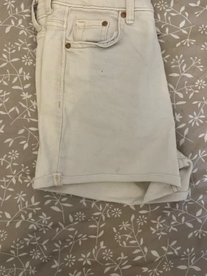 Vita denimshorts med fickor - Säljer ett par vita denimshorts med klassisk femficksdesign och uppvikta benslut. Shortsen har bälteshällor och metallnitar vid fickorna för extra detalj. Perfekta för varma dagar och enkel att matcha med allt. Säljs eftersom de är för små