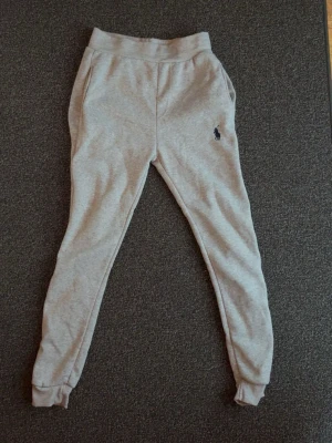 Grå mjukisbyxor från Polo Ralph Lauren - Snygga grå mjukisbyxor från Polo Ralph Lauren med klassisk logga broderad på ena benet. Byxorna har resår i midjan och vid bensluten samt två sidofickor. Perfekta för en chill och avslappnad stil.