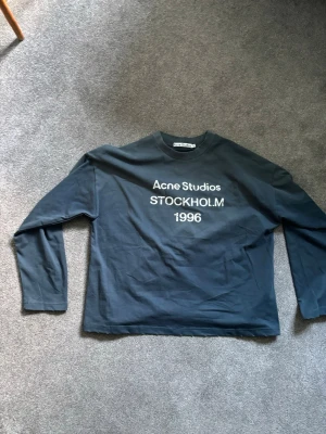 Mörkblå långärmad tröja Acne Studios - Snygg mörkblå långärmad tröja från Acne Studios med vit text 'Acne Studios STOCKHOLM 1996' på bröstet. Tröjan har lite baggy passform, rund halsringning och är gjord i mjuk bomull. Perfekt för en clean och stilren look. PRUTBART!