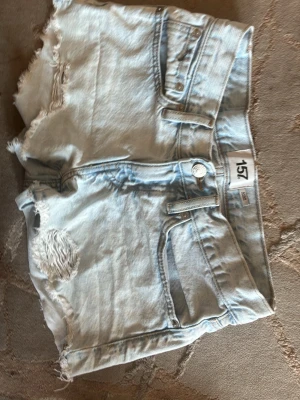 Ljusa jeansshorts från lager 157 - Säljer ett par ljusblå jeansshorts från lager 157 med fransiga kanter och slitna detaljer framtill. Klassisk femficksmodell med knapp och dragkedja. Perfekta för en avslappnad och trendig look i sommar.