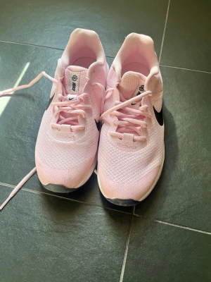 Nike rosa sneakers med svart swoosh - Snygga rosa sneakers från Nike med svart swoosh på sidan. Skorna har en låg profil, mesh-överdel för bra andningsförmåga och vita sulor med lätt struktur. Perfekta för dig som vill ha en sportig och trendig look.