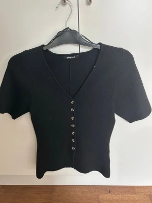 Svart kortärmad kofta Gina Tricot - Stilren svart kofta från Gina Tricot med korta ärmar och v-ringning. Koftan har en figurnära passform och knäpps med små silvriga knappar framtill. Perfekt att matcha med jeans eller kjol för en trendig look.
