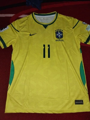 Brasilien matchtröja nummer 11 Nike - Gul Brasilien matchtröja från Nike med nummer 11 på bröstet. Tröjan har gröna detaljer vid krage och sidor, samt det klassiska CBF-emblemet med fem stjärnor broderat på bröstet. Perfekt för dig som älskar fotboll och vill stötta landslaget.