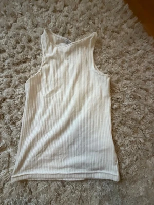Ribbad vit ärmlös topp - Säljer en ribbad vit ärmlös topp med rund halsringning. Materialet är stretchigt och mjukt, perfekt för en avslappnad och trendig look. Passar till jeans eller kjol för en enkel stil.
