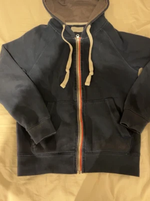 Pier One zip tröja - Mörkblå hoodie från Pier One med dragkedja och beige snören. Insidan av huvan är brun och har ett mjukt tyg. Fickor framtill och en färgglad rand längs dragkedjan ger en cool detalj. Perfekt för en avslappnad stil.