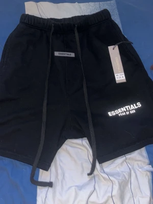 Svarta shorts Essentials Fear of God - Svarta shorts från Essentials Fear of God med elastisk midja och långa snören framtill. Logga tryckt på vänster ben och etikett i midjan. Perfekta för en avslappnad streetwear-look.