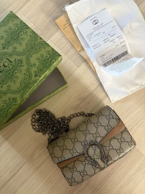  Väska - Säljer en handväska i beige GG Supreme canvas med bruna läderdetaljer och silverfärgad kedja. Väskan har det klassiska GG-mönstret och ett dekorativt spänne framtill. 