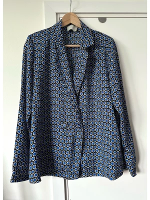 NEO NOIR - blå, svart & vit mönstrad blazer / kavaj - blus, storlek L - Cool mönstrad blazer / kavaj / kavaj - blus från Neo Noir i storlek L. Härligt blommönster i blått, svart och vitt. Tidlös modell med långa ärmar, klassisk krage och knäppning framtill med vackra diskreta klädda knappar. Diskreta tunna inbyggda axelvaddar för bästa passform. Är lika fin att styla som kavaj över topp eller som den är som blus. Finvävt lent satin-material. Mycket gott skick, som ny!