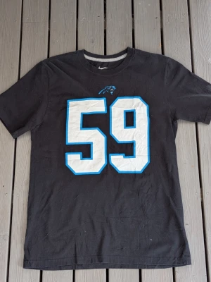 Svart Panthers t-shirt med nummer 59 - Säljer en svart t-shirt med stort vitt nummer 59 och blå kant på framsidan. Ovanför siffran finns Carolina Panthers-loggan tryckt. Perfekt för dig som gillar amerikansk fotboll och vill ha en sportig look.