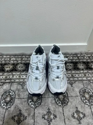  New Balance 530  - Snygga vita New Balance 530 sneakers med silverdetaljer och meshpaneler för extra ventilation. Klassisk låg modell med bekväm sula och tydlig N-logga på sidan. Perfekta för dig som gillar sportig och clean stil.