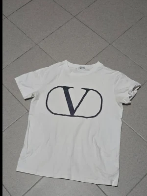 Vit t-shirt med stor V-logga - Vit t-shirt från Valentino med stor svart V-logga på bröstet. Klassisk rund hals och korta ärmar. Tillverkad i mjuk bomull som känns skön mot huden. Perfekt för en stilren och trendig look.