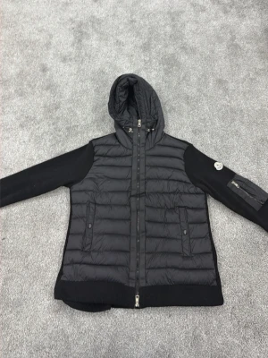 Svart dunjacka med huva från Moncler - Snygg svart dunjacka från Moncler med huva och dragkedja framtill. Jackan har stickade ärmar, quiltad kropp och en ficka med dragkedja på ena ärmen. Moncler-logga på vänster ärm. Perfekt för kalla vinterdagar.