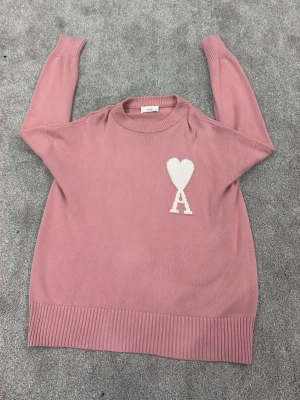 Rosa stickad tröja från Ami - Snygg rosa stickad tröja från Ami med ett vitt hjärta och bokstaven A på bröstet. Tröjan har rund halsringning, ribbade muddar och nederkant. Perfekt för dig som gillar stilrena och trendiga plagg.