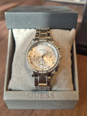 Silverfärgad klocka från Guess - Stilren och glittrig klocka från Guess i silverfärg med länkarmband och stenar runt urtavlan. Urtavlan har flera små visare och tydliga markeringar. Perfekt accessoar för att lyfta din outfit. Det förljer med två bortagana länkar från den