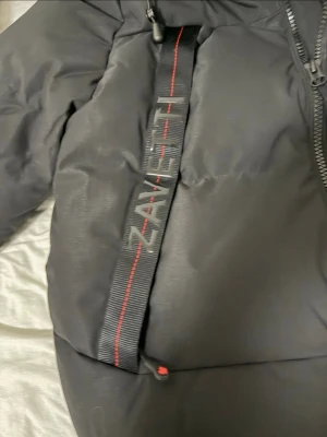 Svart dunjacka från Zavetti Canada - Snygg svart dunjacka från Zavetti Canada med huva och dragkedja framtill. Jackan har detaljer med röda sömmar och logotyp på banden vid bröstet. Perfekt för kalla vinterdagar och riktigt varm tack vare dunfyllningen.