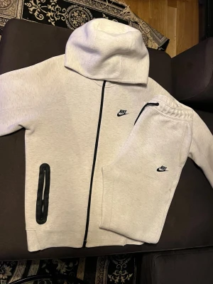 vit Nike tech dress - Säljer ett stilrent vit  loungewear set från Nike med matchande hoodie och byxor. Setet har svarta detaljer, dragkedja på hoodien och elastisk midja på byxorna. Båda plaggen har Nike-logga broderad framtill. Perfekt för chill dagar hemma.