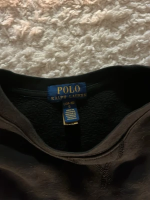 Svart sweatshirt från Polo Ralph Lauren - Klassisk svart sweatshirt från Polo Ralph Lauren med rund halsringning och den ikoniska röda logotypen broderad på bröstet. Tröjan har ribbade muddar vid ärmslut och nederkant, perfekt för en avslappnad och stilren look passar folk runt 170 cm . 